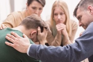 Primer plano de un joven devastado con la cabeza entre las manos y amigos que lo apoyan durante la terapia grupal