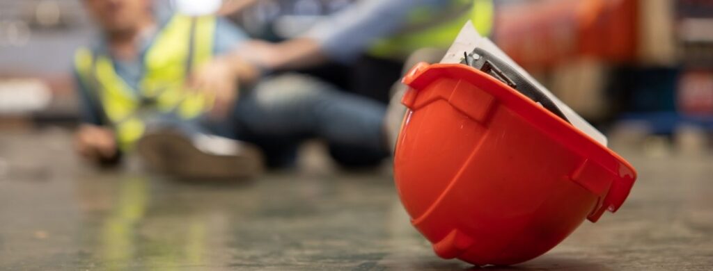 A red hard-hat on the floor with a blurred injured worker in the background. 